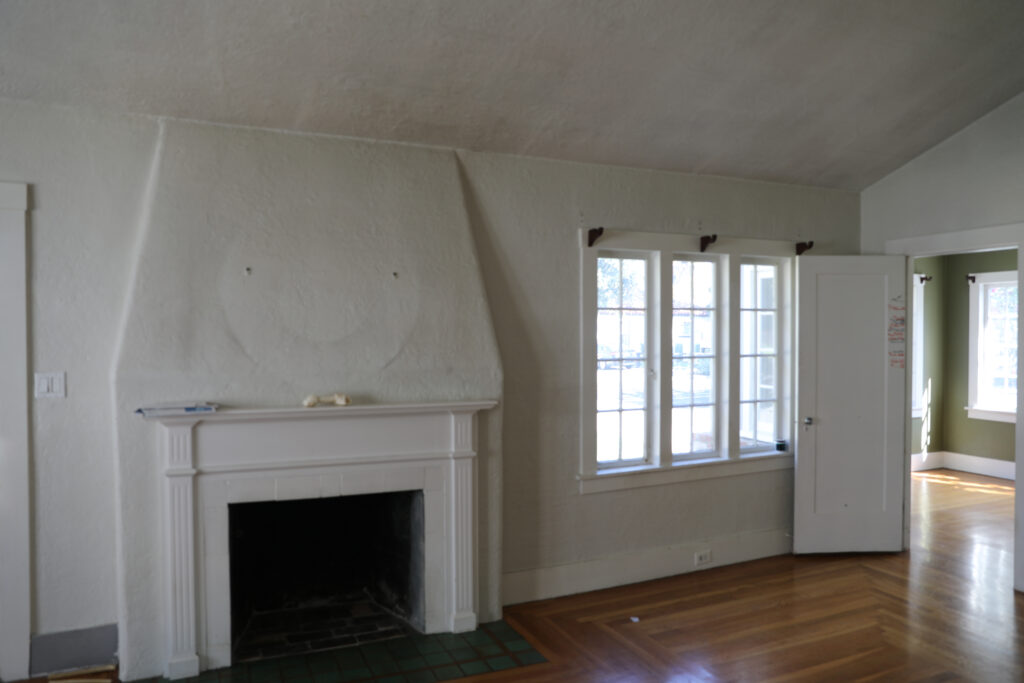 Before renovation living room in older Willow Glen home with worn plaster walls, outdated windows, and a non-functioning fireplace.